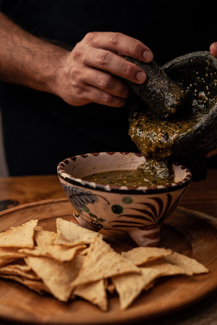 Hands preparing salsa in a traditional mortar, served with nachos on a wooden plate.